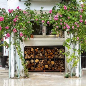 A set of 8 corten steel square log stores with logs inside underneath a window on an outdoor patio with green plants growing either side
