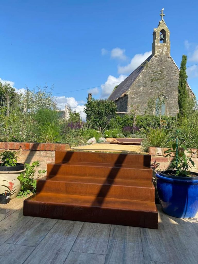 a set of corten steel steps in an outdoor garden with a church in the background