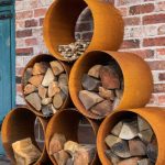 set of 6 corten round log stores with logs and kindling in the top against a brick wall