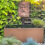 corten steel trough in an outdoor garden with water flowing from a feature with plants growing up a fence behind it